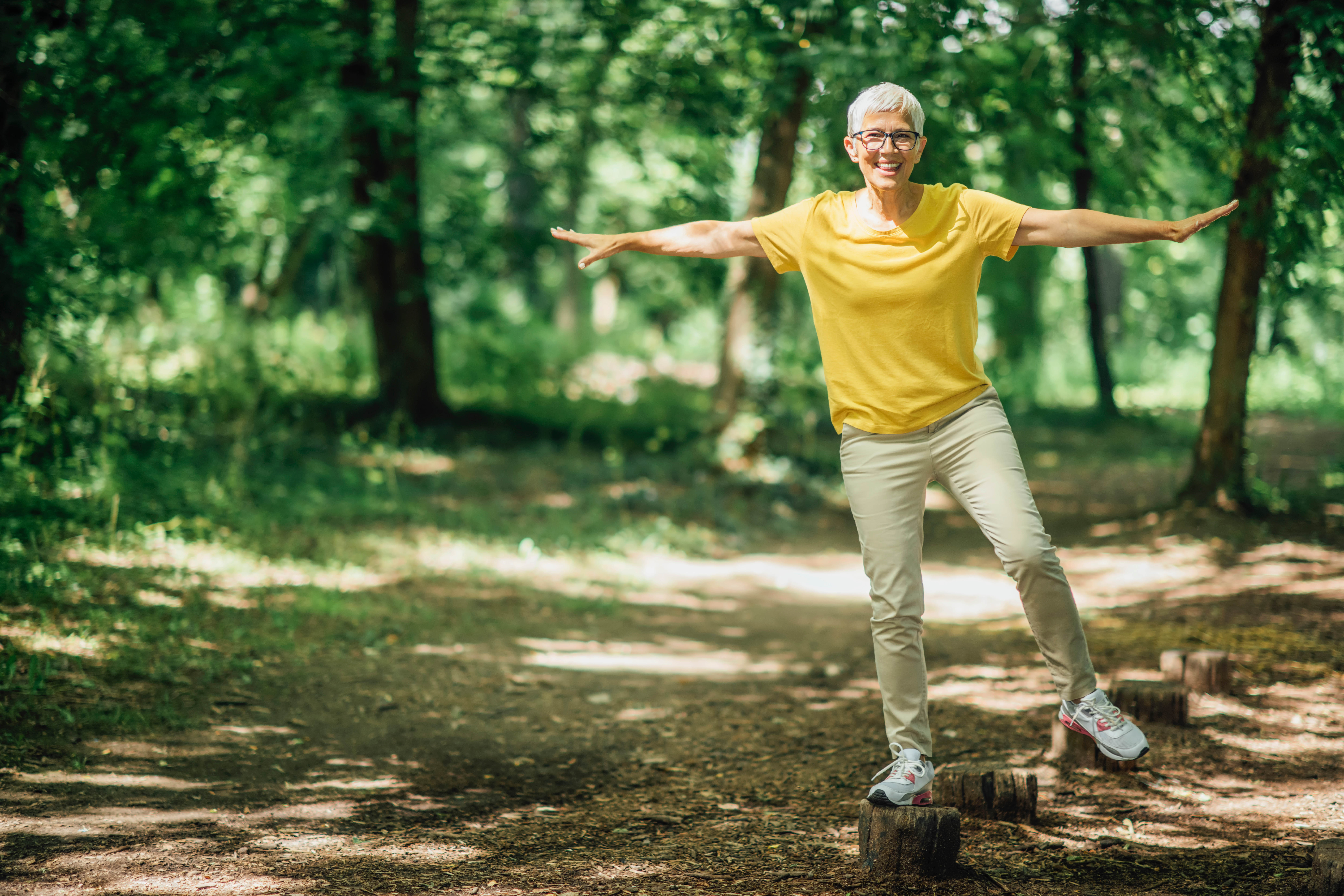 Older woman standing on a tree stump on one foot, doing some balance work outdoors Older woman standing on a tree stump on one foot, doing some balance work outdoors